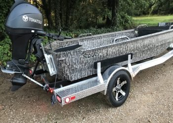 Camo fishing boat on a trailer in front of trees.