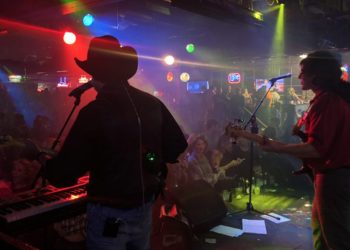 Band with guitars on stage with lights and audience at tables.