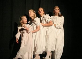 Four young actresses in white nightgowns walk in front of a dark background on stage. 